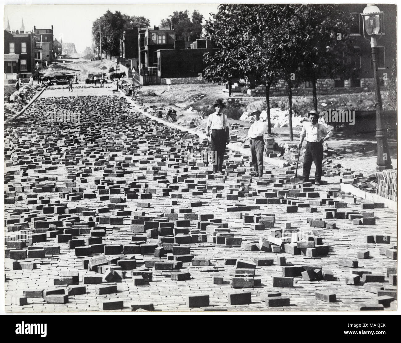 Horizontal, schwarz-weiß Foto zeigt drei Männer Compton mit Ziegelsteinen zu ebnen. Die Männer stehen auf der rechten Seite des Fotos am Rand der Straße und halten eine Schaufel und eine nicht identifizierte. Die Straße führt sie durch die linke Seite des Bildes, und Backsteine sind überall verstreut. Ein typisiertes Hinweis auf der Rückseite der Drucken, möglicherweise eine Ausstellung Bildunterschrift, lautet: 'Straße ebnet. 1907. Pflasterung Straßen mit Backsteinen war eine lange und mühsame Arbeit, bevor noch mehr moderne Methoden der Oberflächenbehandlung entwickelt wurden. Regen und Schnee machte diese Straße sehr gefährlich und An schlechten Tagen spezielle Stockfoto