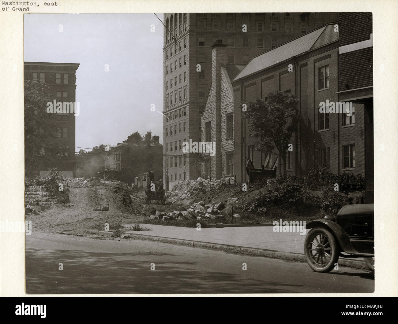 Horizontal, Schwarz und Weiß Foto, Bauarbeiten auf der Washington Avenue cut-off, östlich von Grand Avenue. Teil einer niedrigen Mauer aus Stein an der Grenze zu den Bürgersteig auf der Washington Avenue wurde abgerissen worden. Ein Mann stand auf einem Wagen gesehen Schaufeln ein Haufen Schmutz hinter der Wand. Washington Avenue verläuft im Vordergrund, und mehrere Stein und Ziegel können Gebäude im Hintergrund gesehen werden. Das vordere Ende einer geparkten Auto kann an der rechten Seite gesehen werden. Titel: Arbeiter beim Bau der Washington Avenue cut-off, östlich von Grand Avenue. . 1916. Holt, Charles Cleme Stockfoto