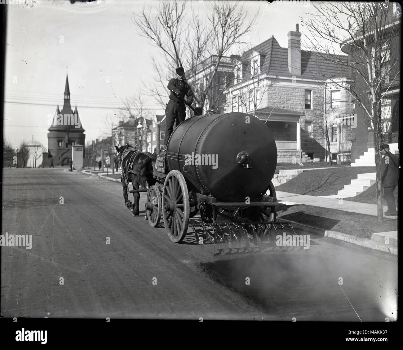 Horizontal, schwarz-weiß Foto eines Pferdes - tanklaster Öl sprühen auf einer Straße mit Wohnhäusern. Es ist ein Fahrer auf der Truck Operating einen Hebel und mit Blick auf den Betrachter. Es gibt Häuser entlang der rechten Seite der Straße sichtbar, und einen eigenen Eingang von der Straße Tor ist im Hintergrund sichtbar. Titel: Straße beölung Lkw bei der Arbeit an der Washington Hotel in der Nähe der Eingang vom Union Boulevard. . Ca. 1905. Holt, Charles Clement, 1866-1925 Stockfoto