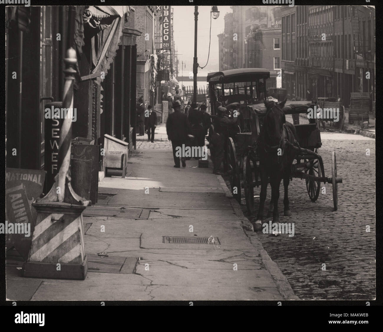 Horizontal, schwarz-weiß Foto mit Blick auf die Straße von der Market Street. Der bürgersteig ist mit Store Fronten und eine gestreifte barber Pole ist an der Vorderseite des Foto links gesäumt. Eine von Pferden gezogene Wagen und andere unattached Karren und Wagen sind gegen den Bürgersteig, wo die Fußgänger Spaziergang entlang geparkt. Titel: Markt (Block 200 nach Osten). . Zwischen ca. 1800 und ca. 1900. Swekosky, William G., 1895-1964 Stockfoto