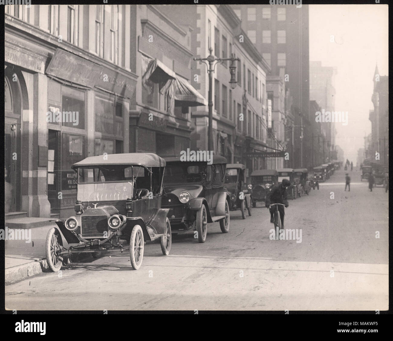 Horizontal, schwarz-weiß Foto mit Blick auf die Straße von der Achten Straße. Eine Reihe von Automobilen Linie der linken Seite der Straße vor einer Reihe von gewerblichen Gebäuden. Ein Mann fährt mit dem Fahrrad in der Mitte der Straße. Titel: Eighth Street Block 100. . Zwischen ca. 1800 und ca. 1900. Swekosky, William G., 1895-1964 Stockfoto