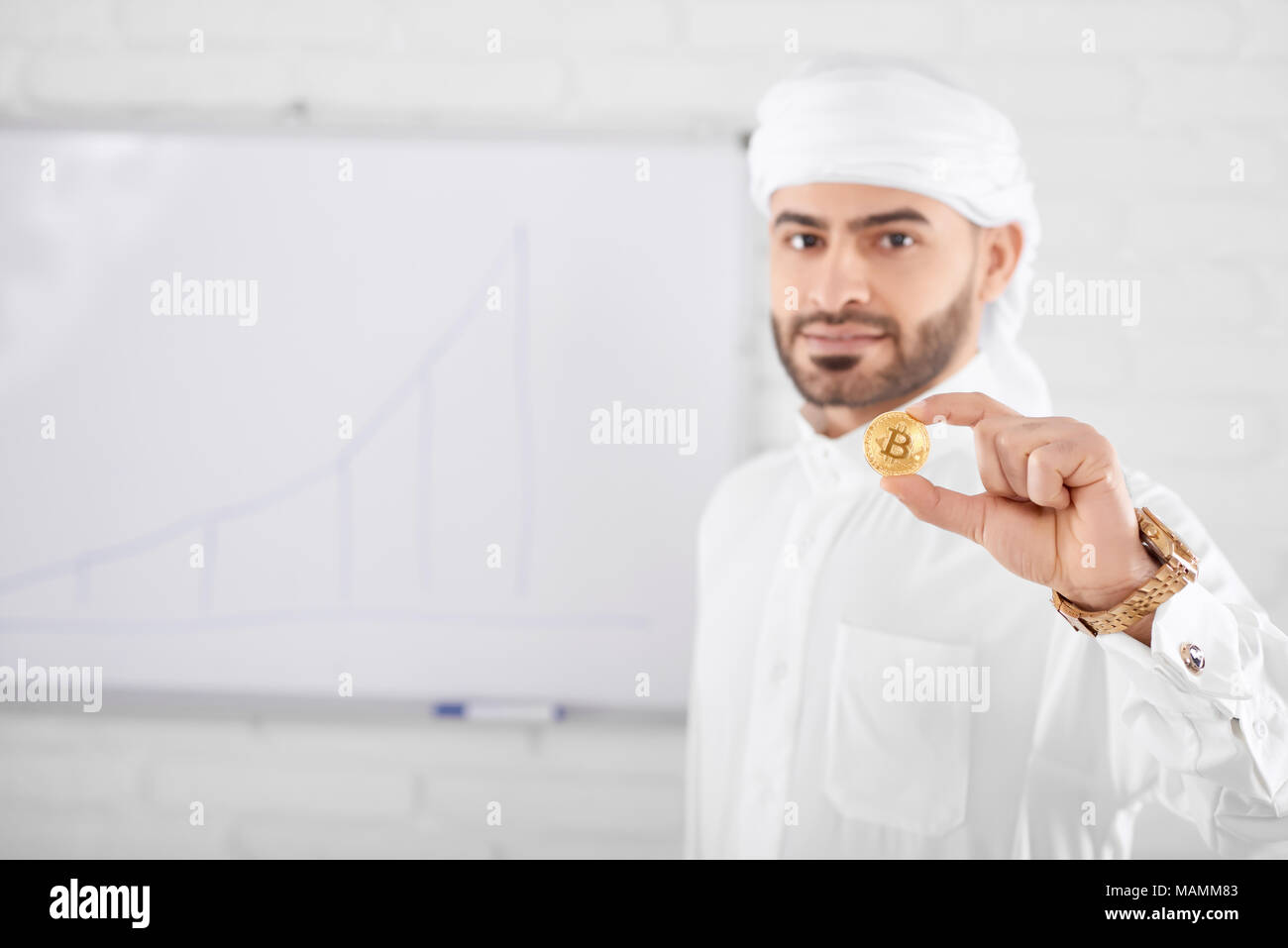 Wohlhabend aussehenden muslimischer männlicher Modell in traditionelle islamische Kleidung und Goldene bitcoin vor der Tafel an der Wand. Selektiver Fokus, unscharfen Hintergrund, horizontal gedreht. Konzept der cryptocurrency. Stockfoto