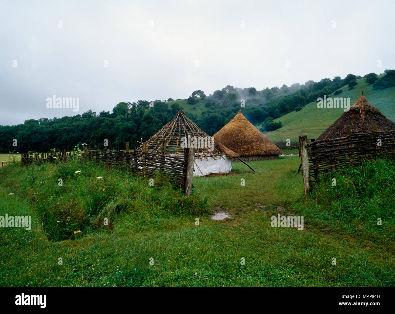 Archäologische Rekonstruktionen der Eisenzeit rundhäuser durch die NE-Eingang eines mit einem Graben und umzäunten Gehege an butser Ancient Farm, UK gesehen. Stockfoto