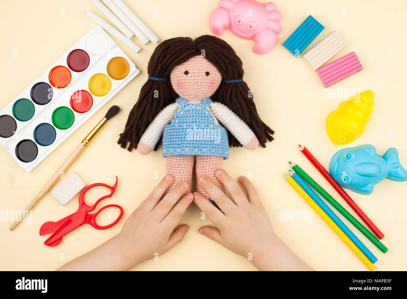 Das Kind sitzt am Tisch mit Objekten für Kreativität, Zeichnung und Hobbys, hält eine gestricken Puppe in den Händen, eine Sicht von oben Stockfoto