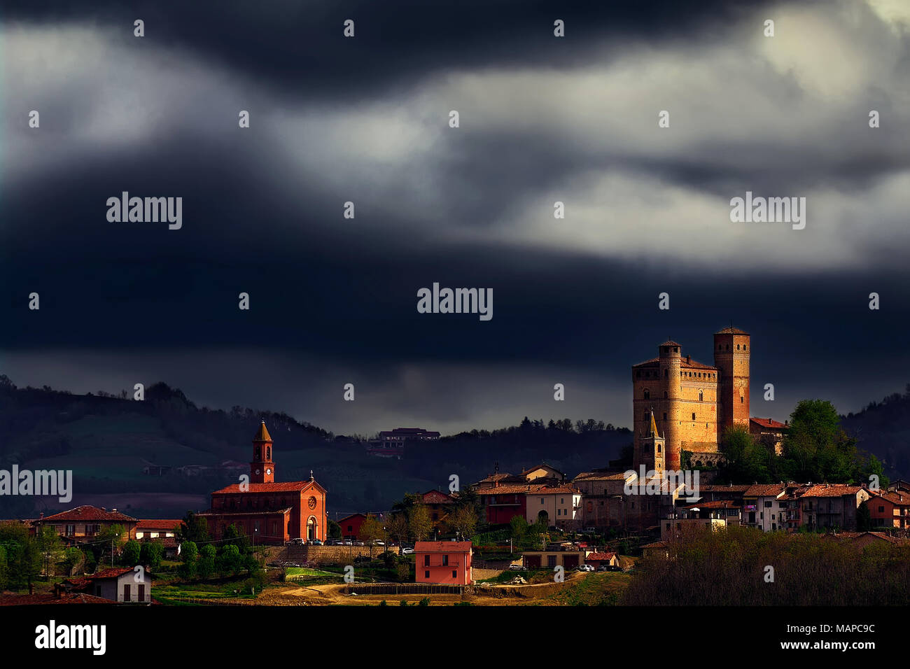 Ein Strahl der Sonne beleuchtet das Schloss und die Kirche von Serralunga d'Alba unter einem stürmischen Himmel. Wir sind in der Mitte der Produktionsbereich des Barolo. Stockfoto