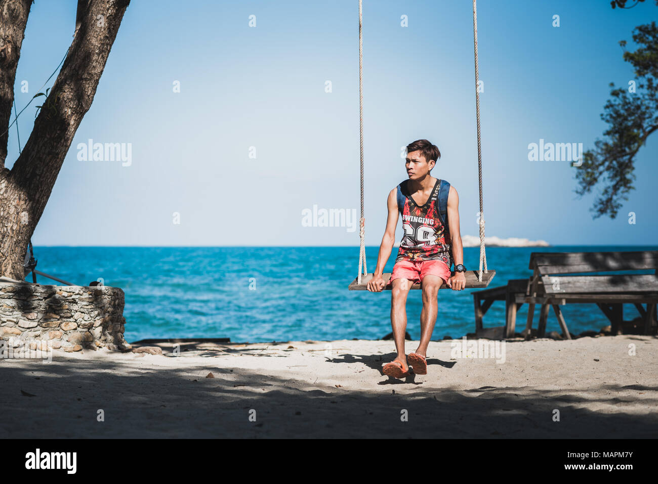 Junger Mann im oberen Tank sitzt auf einem Seil schwingen auf sandigen Ufer gegen den blauen Meer und den klaren Himmel. Ko Samet (Koh Samed), Thailand Stockfoto