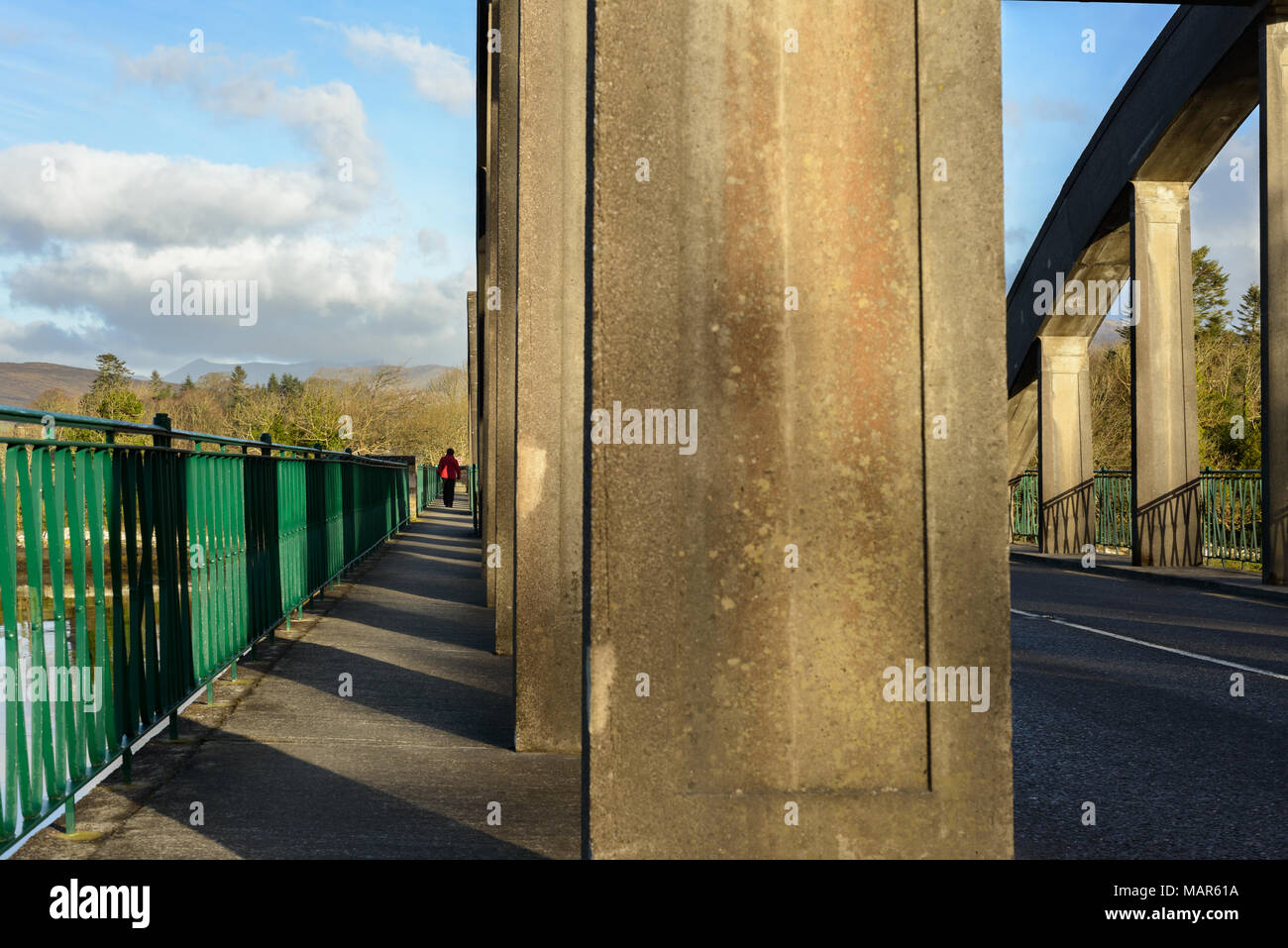 Einsame Person in roter Jacke zu Fuß auf die Brücke entlang Zement Spalten, die Hängebrücke, der Klang, Kenmare. Früh sonnig irischen Morgen. Stockfoto