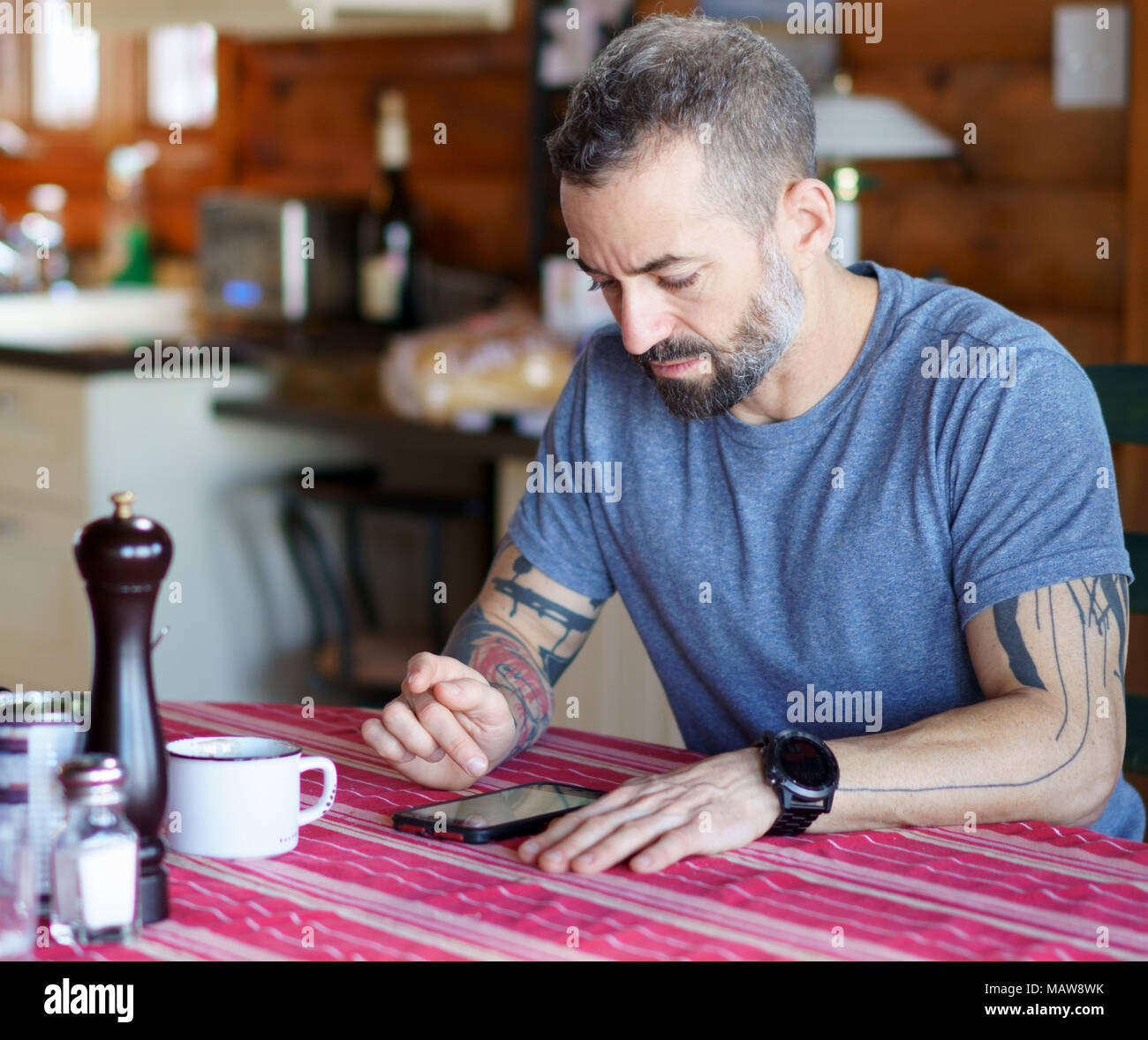 Von Mitte 30 Bart und tätowierte Mann an seinem Smartphone am Küchentisch mit einer roten Tischdecke und trägt ein blaues T-Shirt und ein großes Ansehen. Stockfoto
