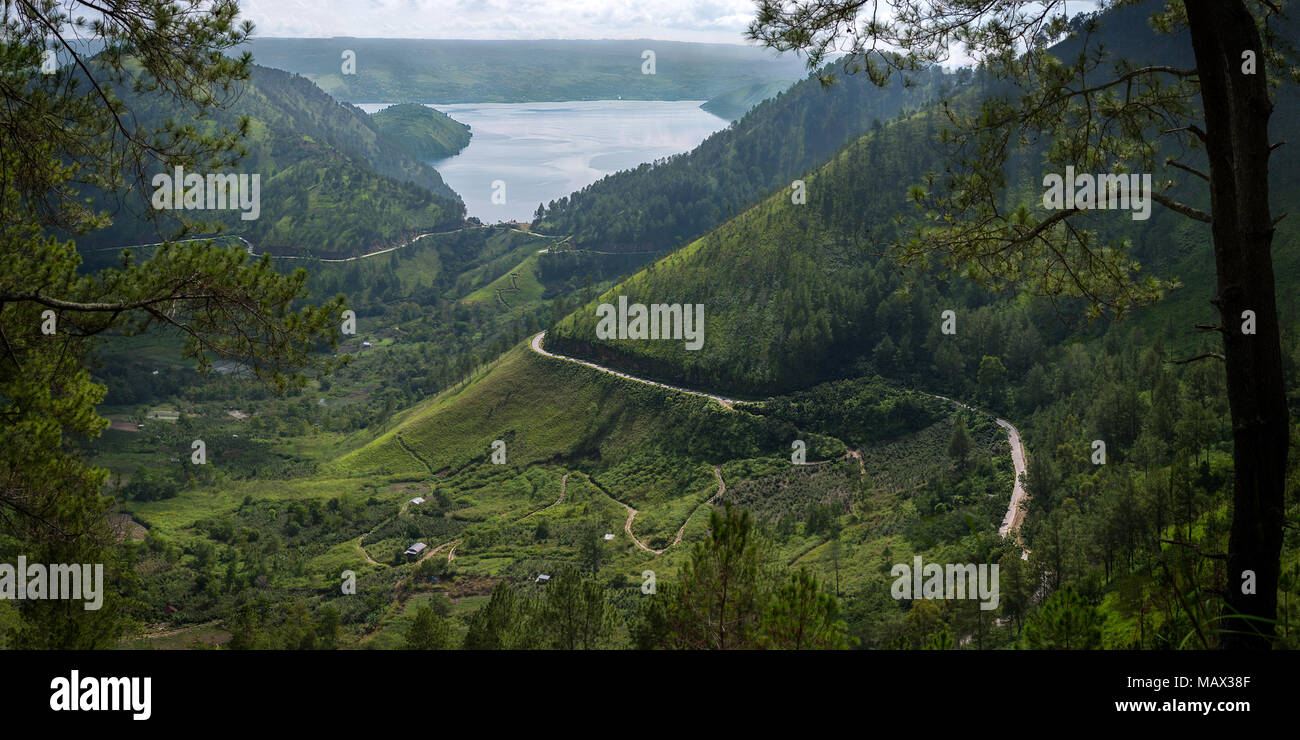 Eine dünne Straßen schneiden sich in den vulkanischen Hügeln rund um Lake Toba Winde hinunter zum See gesehen von Westen durch die umliegenden Bäume. Sumatra, Indones Stockfoto