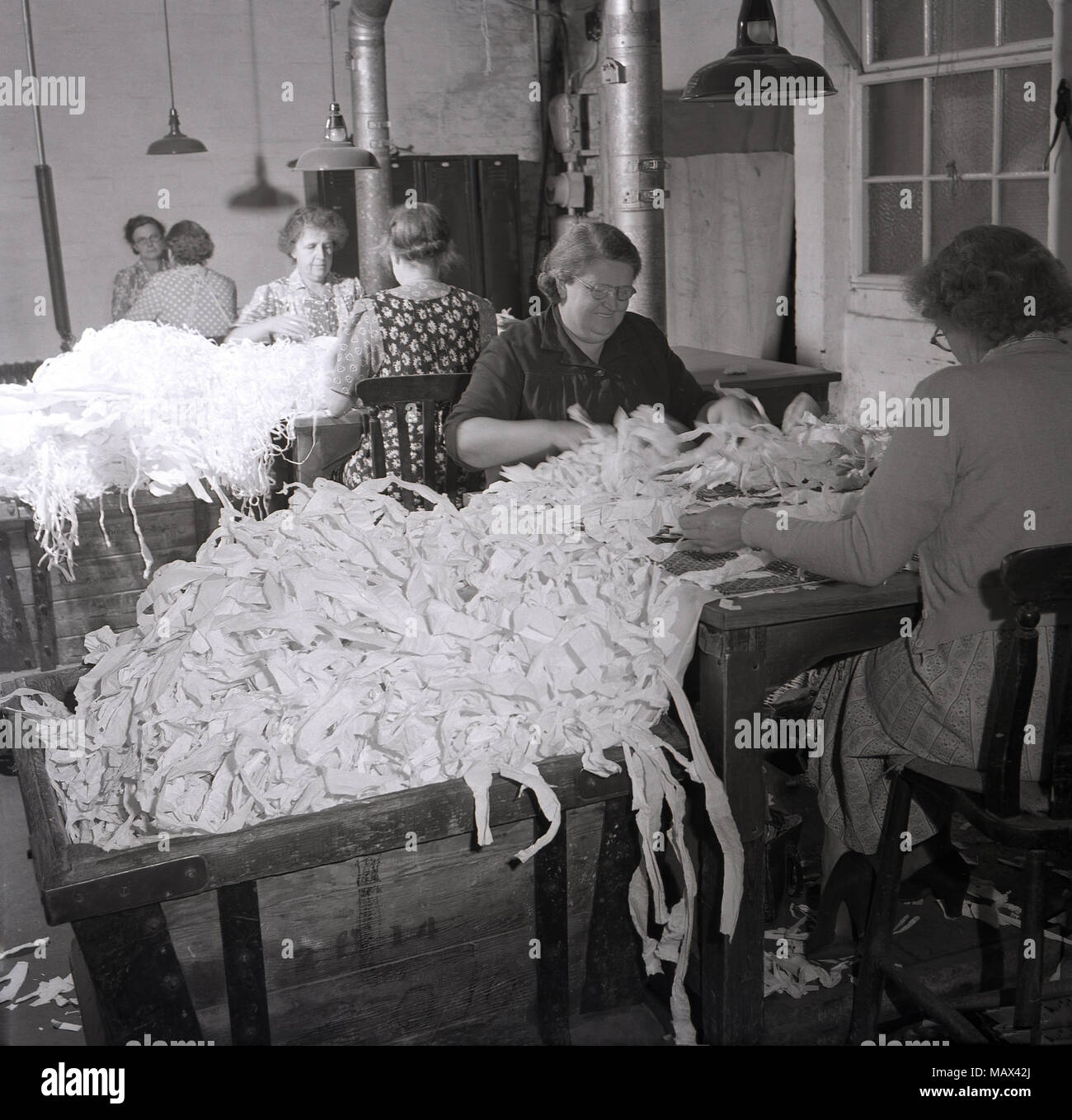 1950er Jahre, historische Frauen, die in einer kleinen Baumwollmühle arbeiten, die sich an Bänken gegenübersitzen und große Bündel von Überresten oder Materialabschnitten aussortieren, möglicherweise Baumwolle, England, Großbritannien. Stockfoto