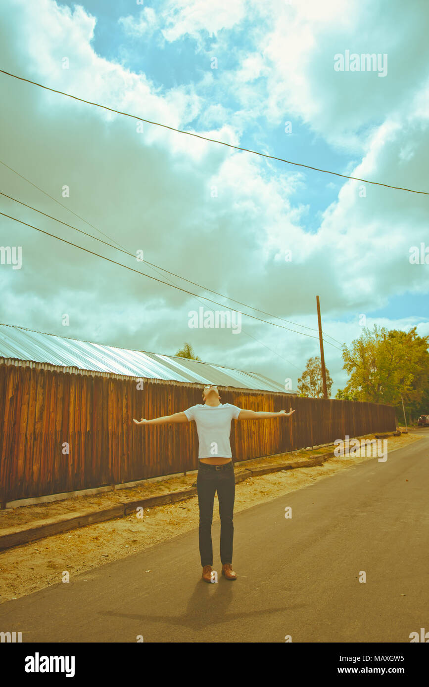Einem Kaukasischen jungen Mann stand auf einer kleinen Landstraße, bis hin zu einem bewölkten Himmel mit seinen Armen Blick; eine geistliche Porträt Konzept. Stockfoto