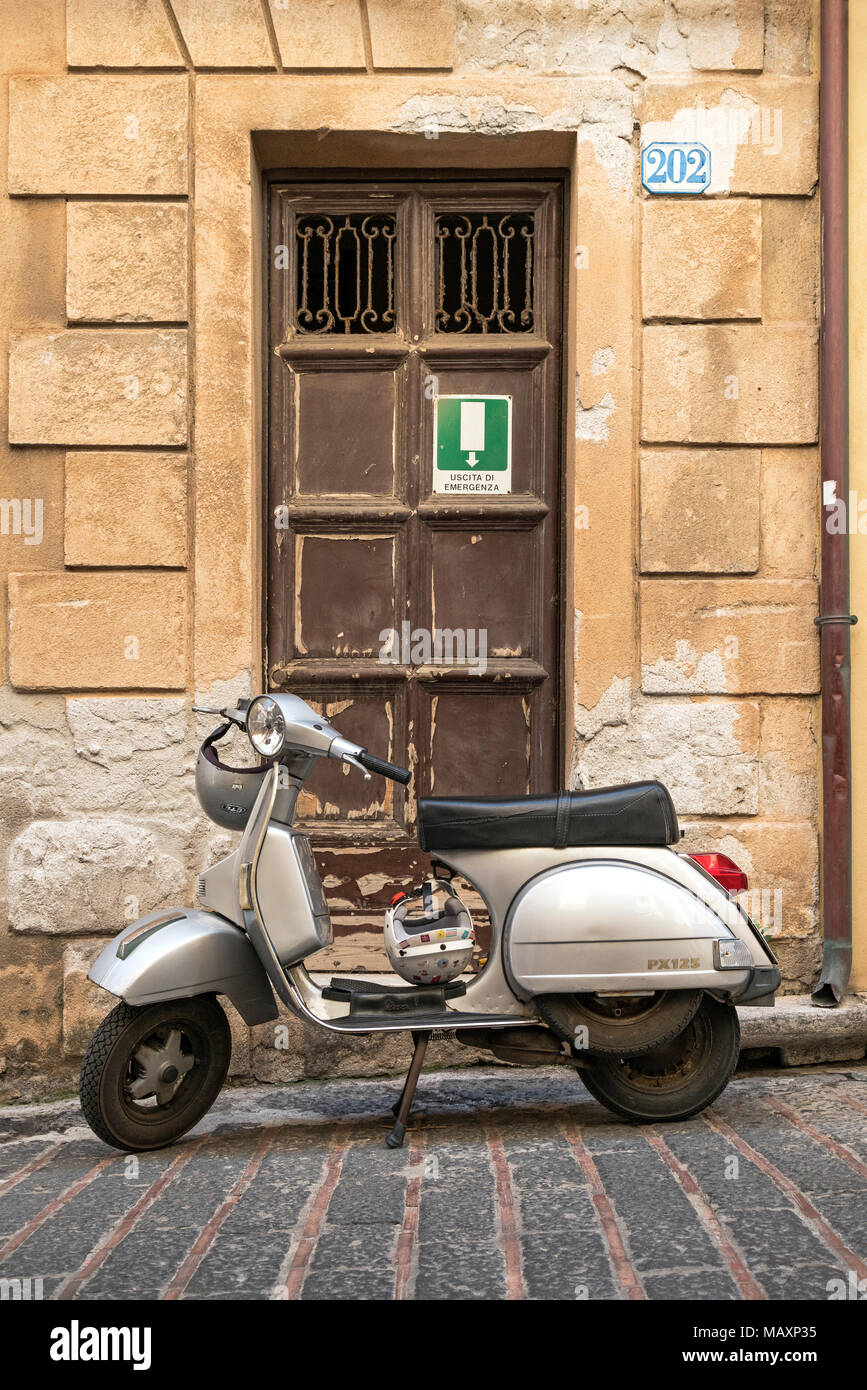 Eine silberne Vespa PX 125 neben eine alte Tür mit Notausgang Schild auf den Straßen von Cefalu in Sizilien geparkt. Stockfoto
