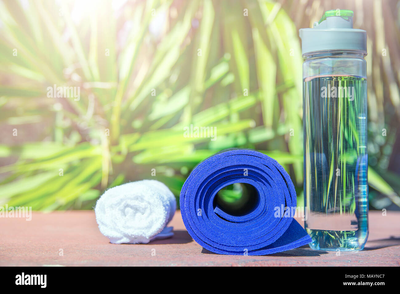 Blau Yogamatte Flasche mit Wasser Weiß Handtuch auf Grün Palme Natur Hintergrund gerollt. Hellen Mittag Sonnenlicht. Entspannung Sommer Meditation Fitne Stockfoto
