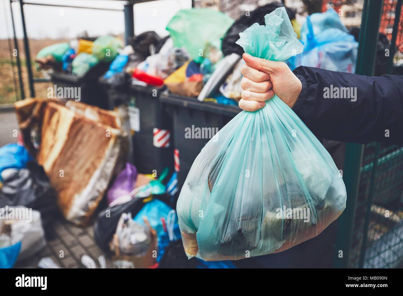 Hand mit Müll gegen den vollen Mülleimer mit Müllsäcke überlaufen auf dem Bürgersteig. Stockfoto