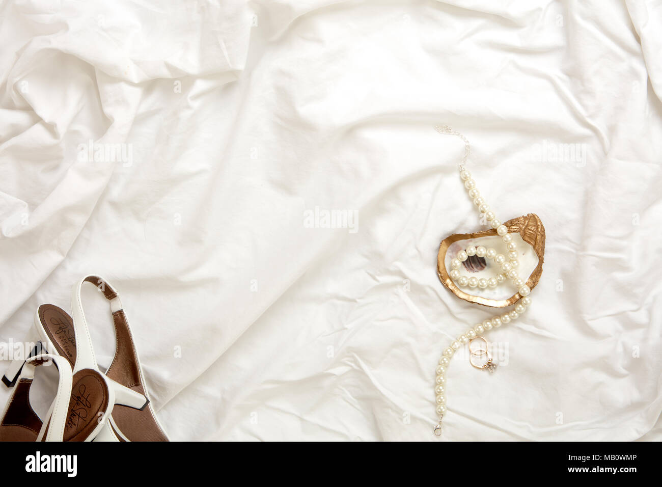 Gold oyster Schmuck Gericht mit Perlenkette, weiß Heels Schuhe und gold Engagement Ring und gold Hochzeit Band auf einem weißen Stoff Hintergrund Stockfoto