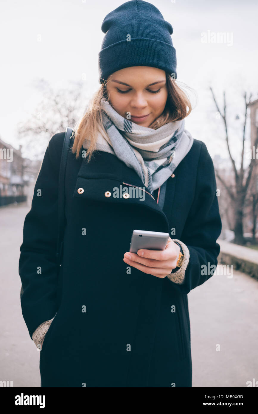 Vorderansicht des lächelnden jungen Frau an der Straße stehen und im  Smartphone. Stilvolle Mädchen mit schwarzen Mantel, blau Schal und Mütze  Hut in Win Stockfotografie - Alamy