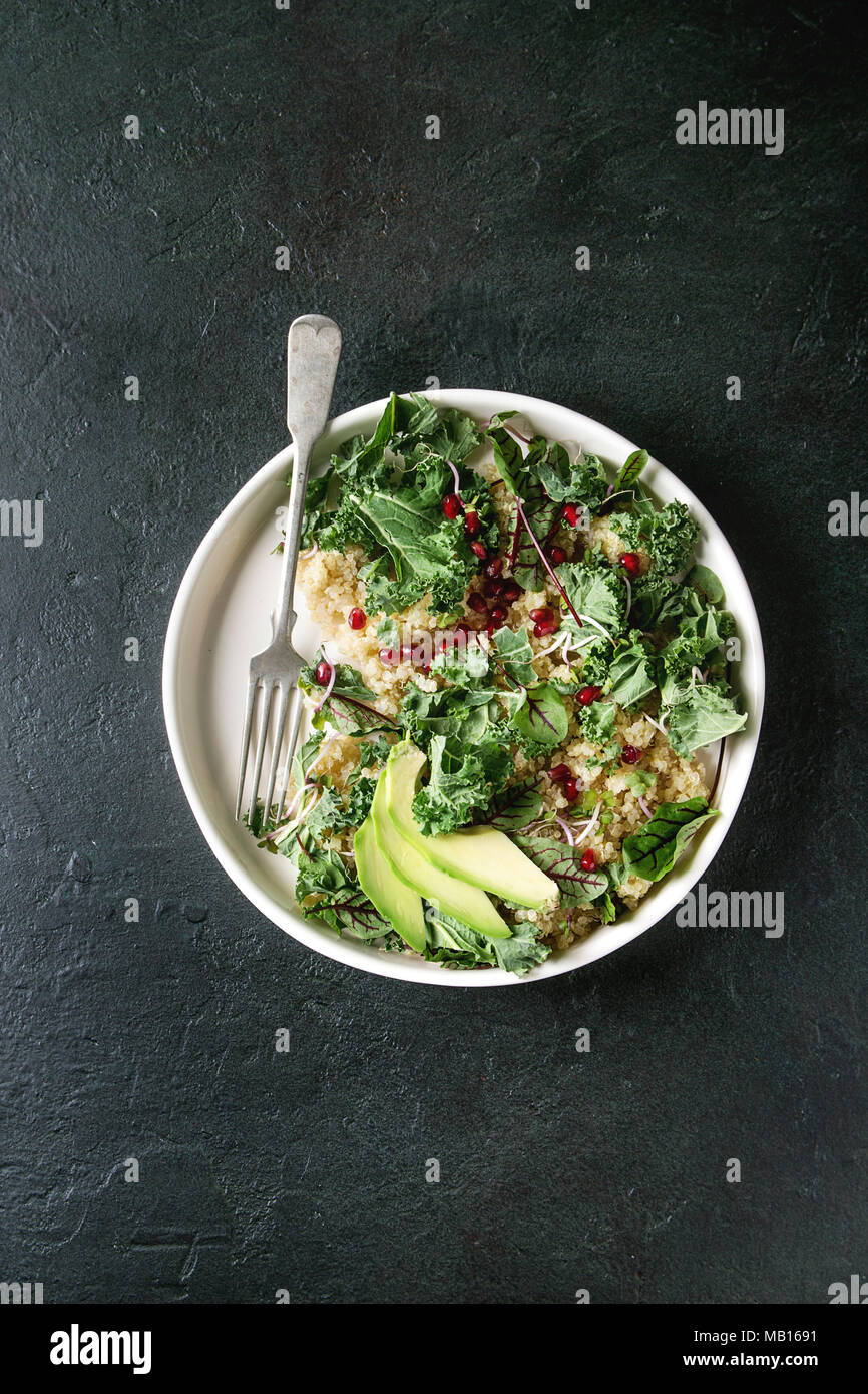 Vegan quinoa Salat mit Grünkohl, junge Rote Bete Blätter, Granat Samen, in Scheiben geschnittenen Avocado in weißer Teller mit Gabel über dunkle Textur Hintergrund. Ansicht von oben, Spa Stockfoto