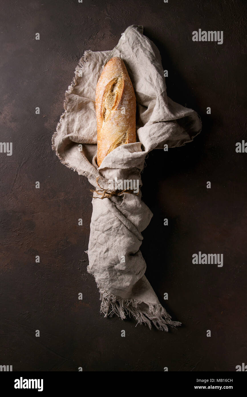 Laib geschnitten frisch gebackene artisan Baguette im Leinen über Dunkelbraun Textur Hintergrund gewickelt. Ansicht von oben, kopieren. Stockfoto