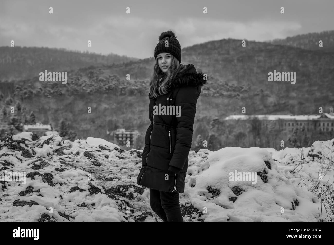 Recht jugendlich Mädchen im Schnee, Kamera, Schwarz und Weiß. Schönen kaukasischen Jugendmädchen in schwarzen Mantel, schneebedeckten Berge im Hintergrund. Stockfoto