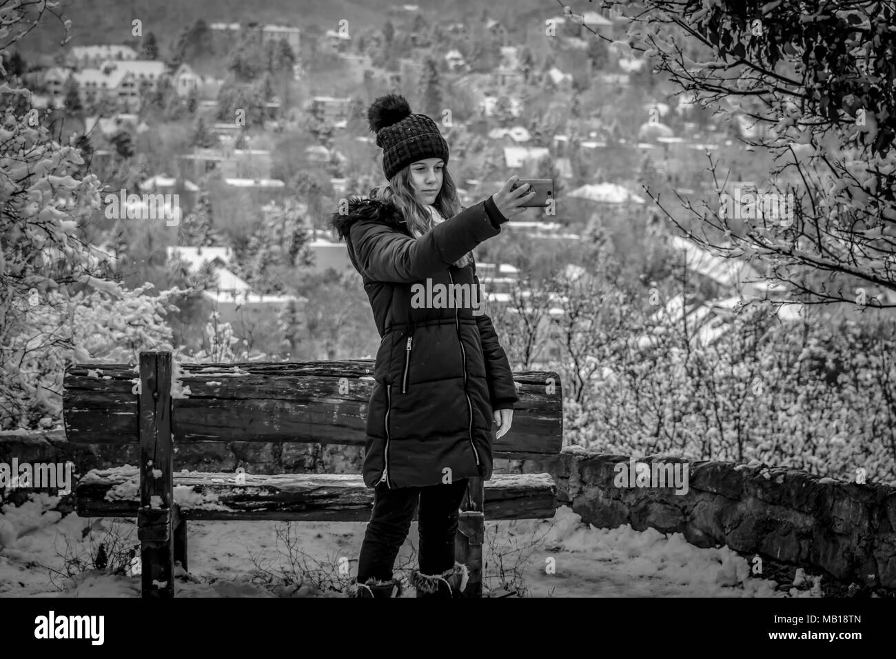Recht jugendlich Mädchen im Schnee, nimmt selfie mit Telefon, Schwarz und Weiß. Schönen kaukasischen Jugendmädchen in schwarzen Mantel, verschwommenes verschneite Stadt im Hintergrund. Stockfoto