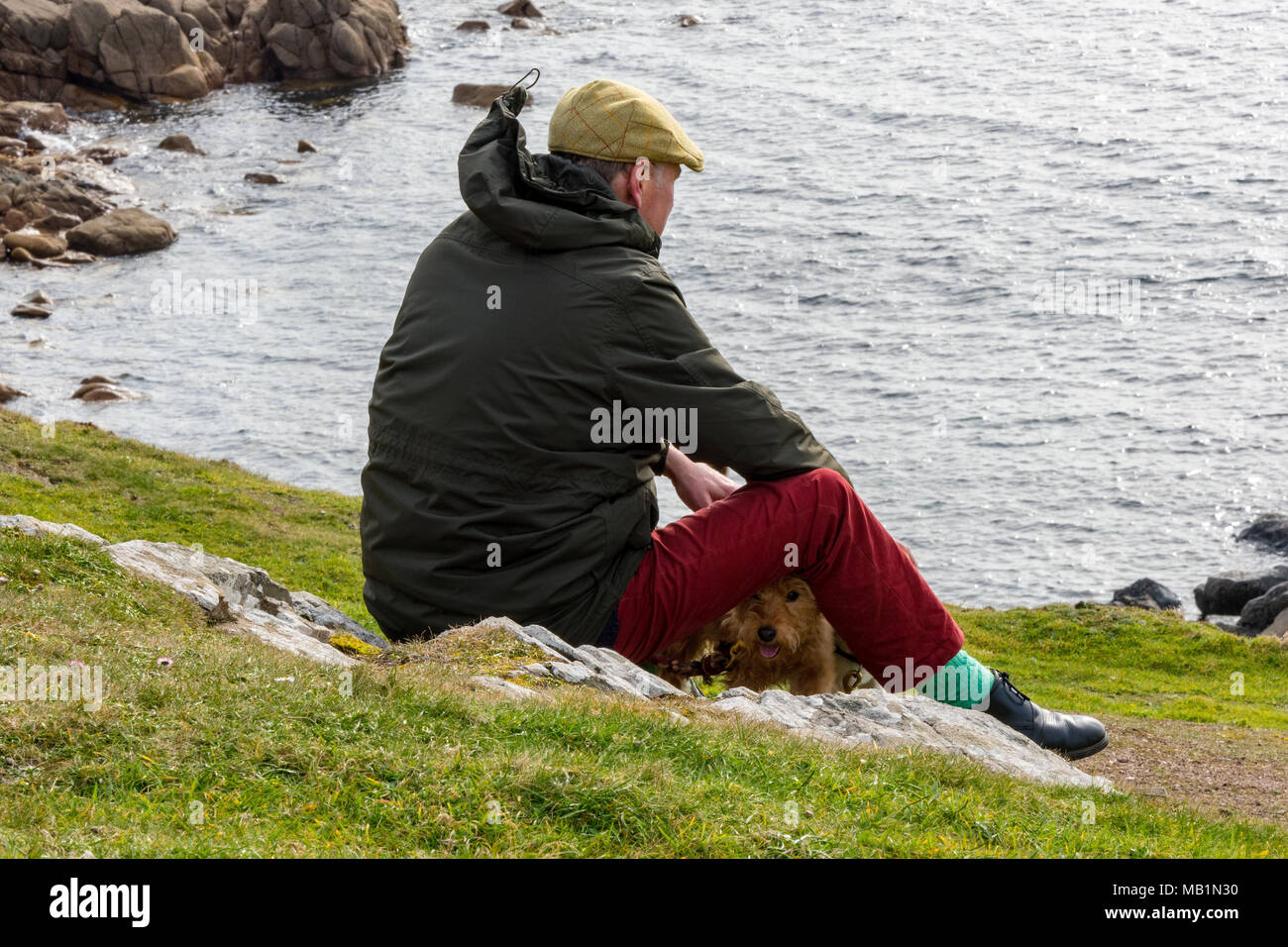 Ein Mann, gekleidet in einem flachen Kappe und Winterjacke sitzt auf der Spitze der Klippen mit einem kleinen Terrier Hund aus zu sehen. Denken gehen der Hund. Stockfoto