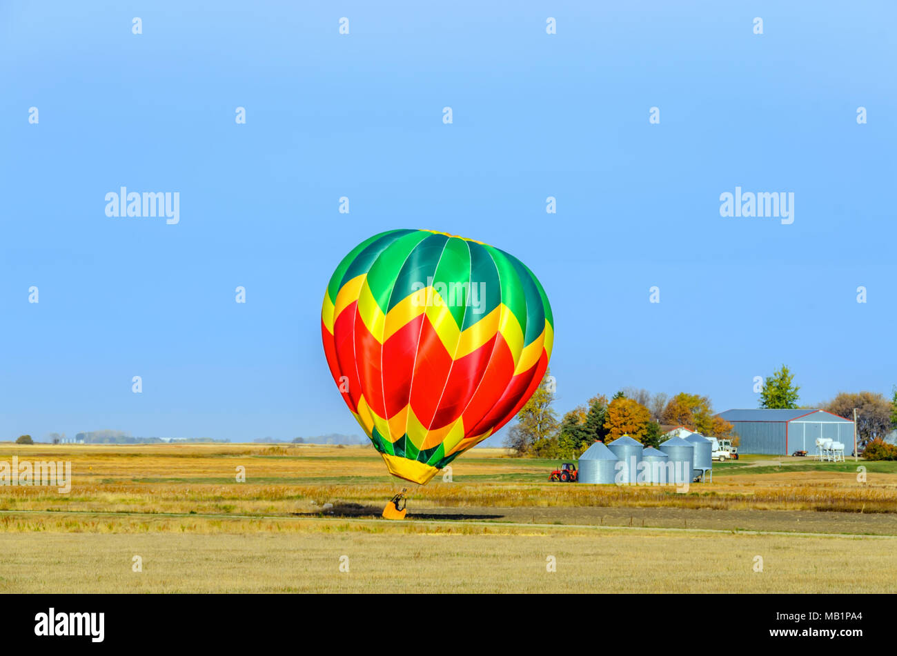 Eine mehrfarbige Ballon mit einem Korb, eine Flamme von Feuer und die Silhouette eines Pilot fliegt in den blauen Himmel über einem gelben Feld an einem Sommertag, ein tra Stockfoto