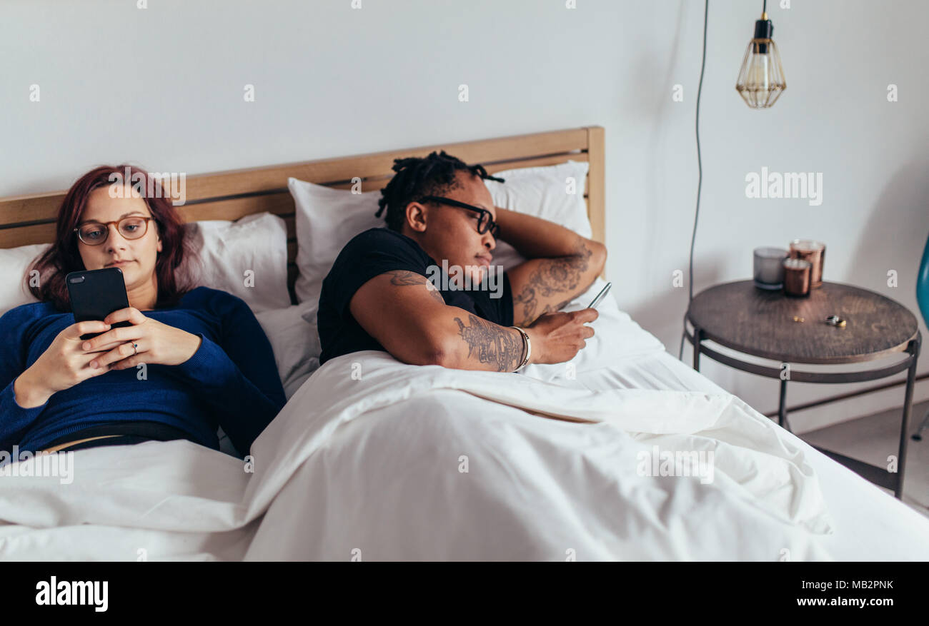 Junge gemischtrassiges Paar mit ihren Handys im Bett ignorieren einander. Mann und Frau beschäftigt mit ihren Handys im Bett. Stockfoto