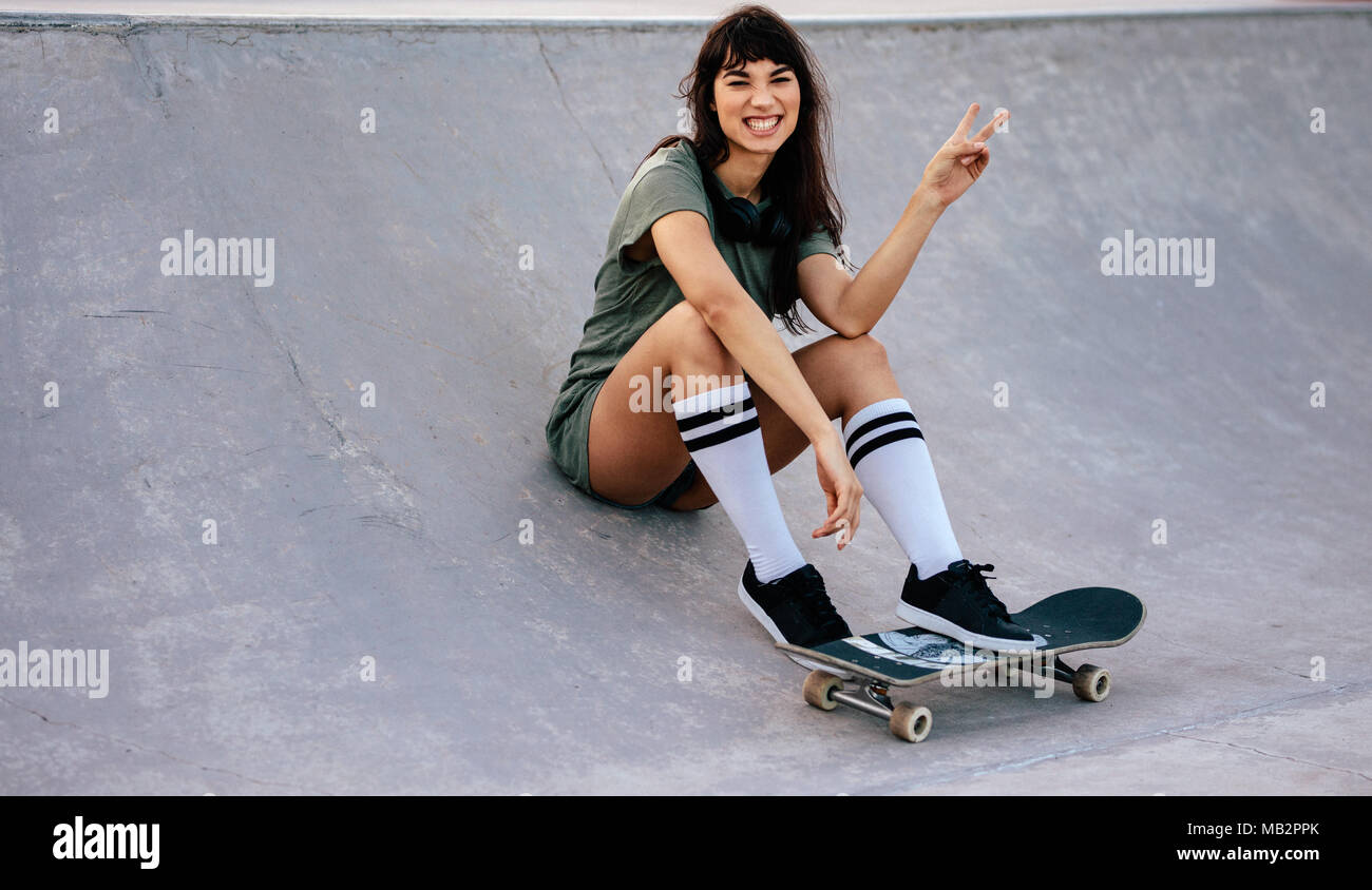 Porträt der schönen jungen Frau an Skate park Rampe sitzend mit einem Skateboard Lächeln und einen Frieden Hand unterzeichnen. Stockfoto