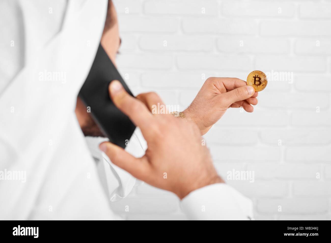 Ein arabischer Mann hält Golden bitcoin in der einen Hand und die Gespräche über das Mobiltelefon mit einer anderen. Er trägt weiße traditionellen muslimischen tragen. Close-up war auf dem Studio Hintergrund. Stockfoto