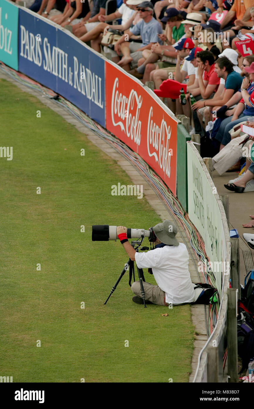 Ein professioneller Fotograf mit einem Teleobjektiv von Canon, montiert auf einem Stativ bei einem Kricketspiel sitzen auf dem in Feld vor der Werbetafel. Stockfoto