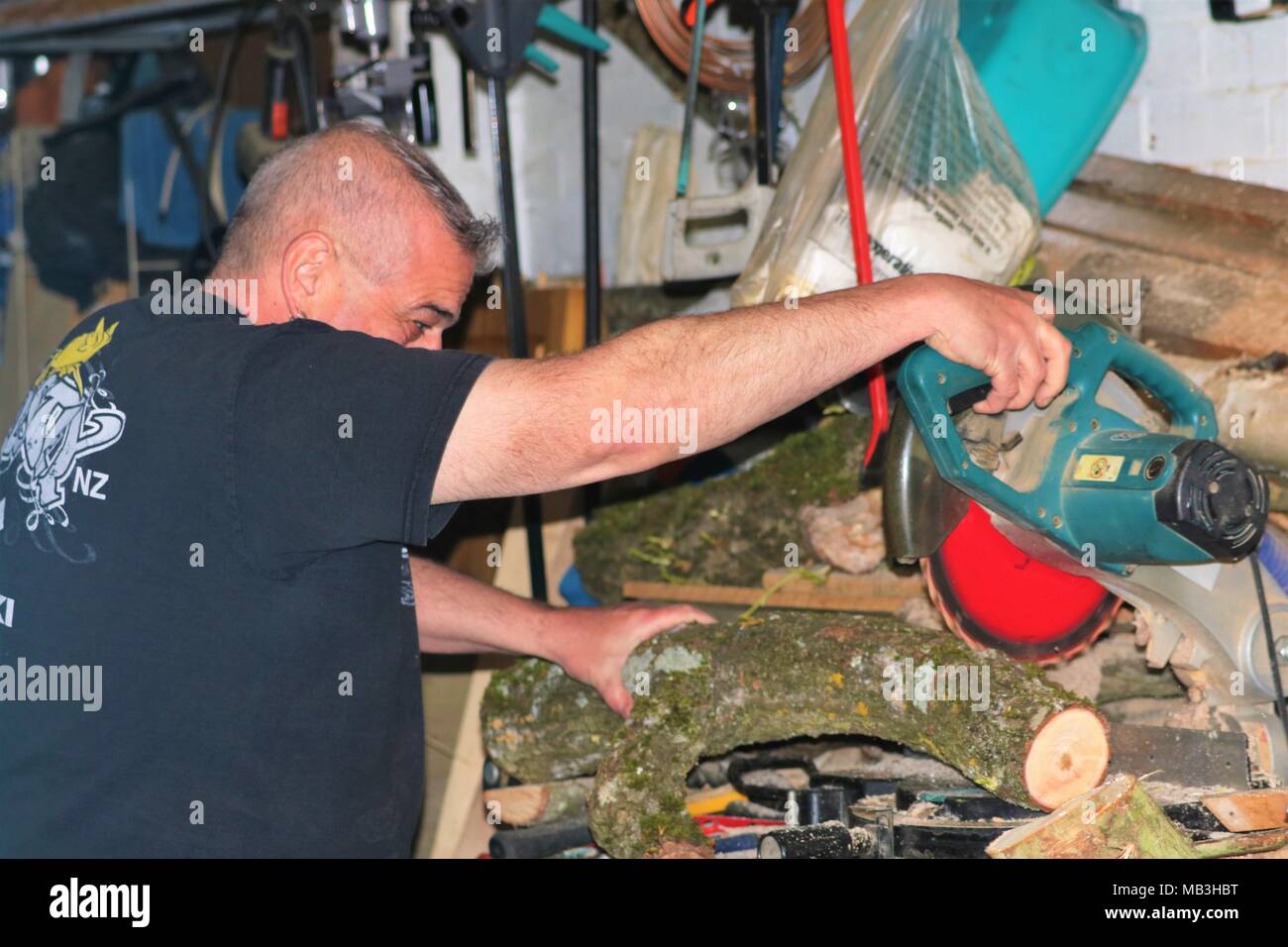 Im mittleren Alter Mann zerschneiden ein Protokoll mit einer Gehrungssäge/sah in einen übervollen Garage hacken und tragen nicht alle Sicherheitseinrichtungen Stockfoto