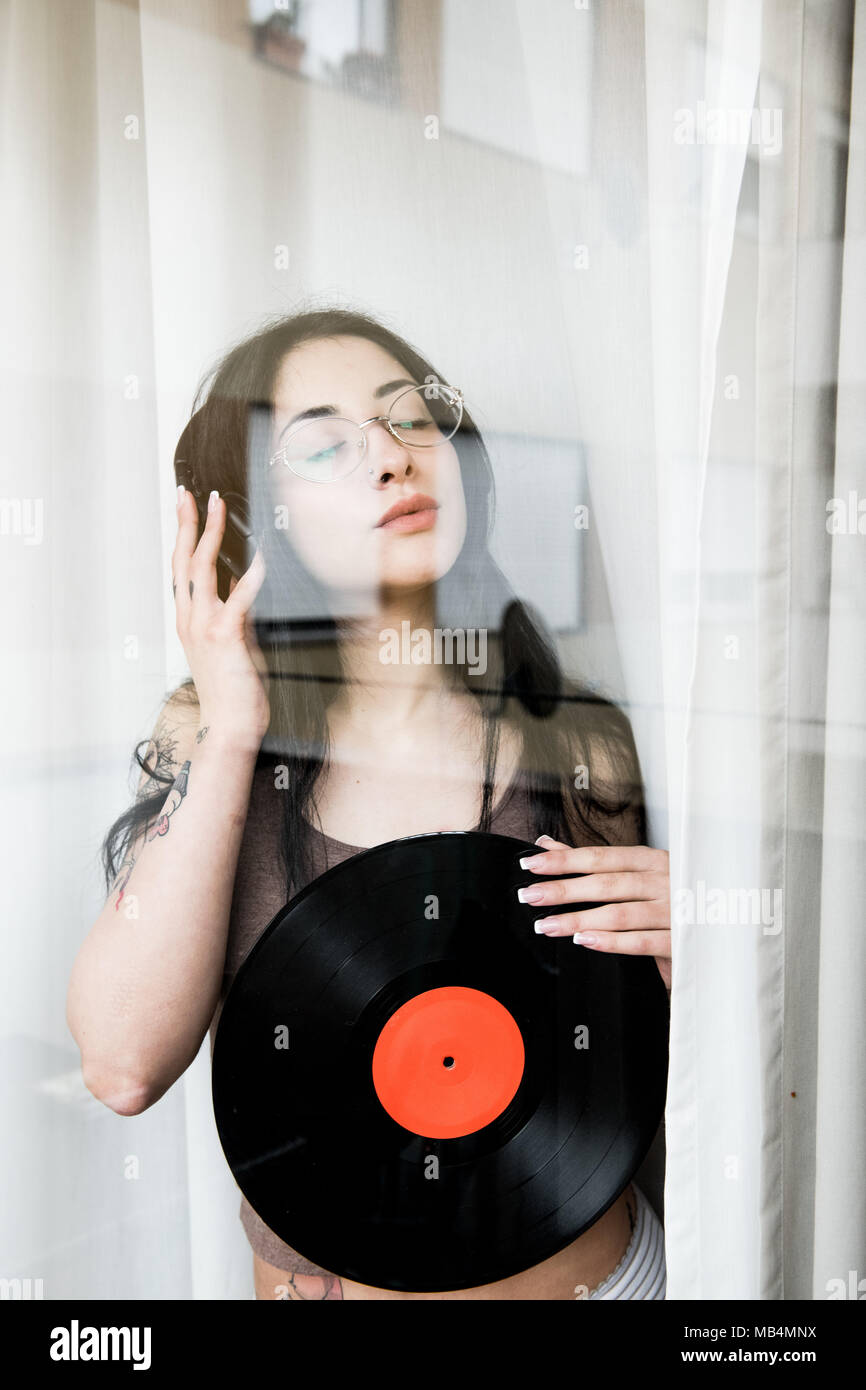 Hübsches Mädchen weibliche Teenager hinter Fenster Musik hören mit Kopfhörern und Schallplatte an Kamera suchen Stockfoto