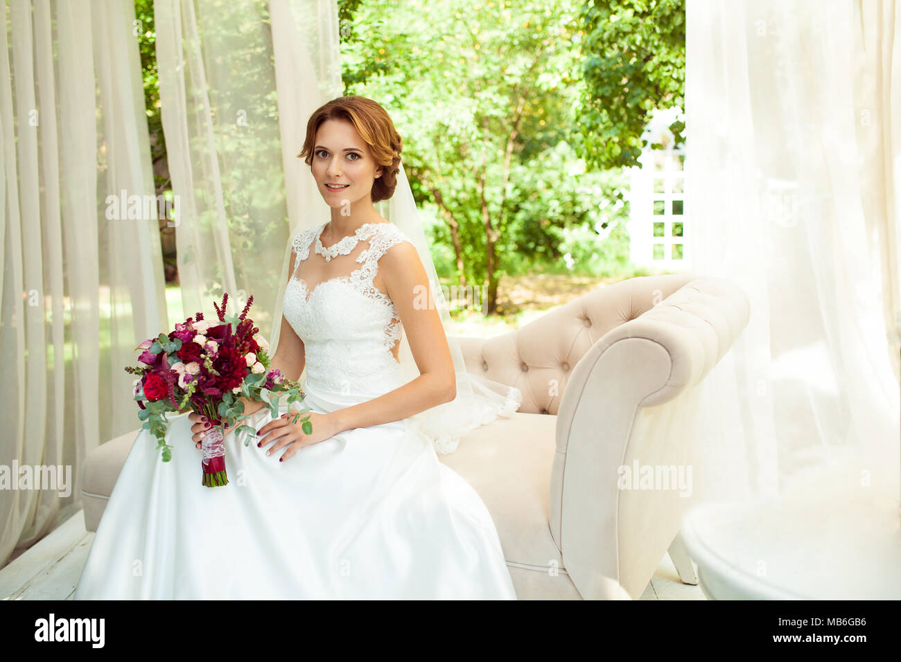 Attraktive Braut mit Blumenstrauß Blume tragen Hochzeit weiß Kleid sitzt auf beige Sofa in Park Stockfoto
