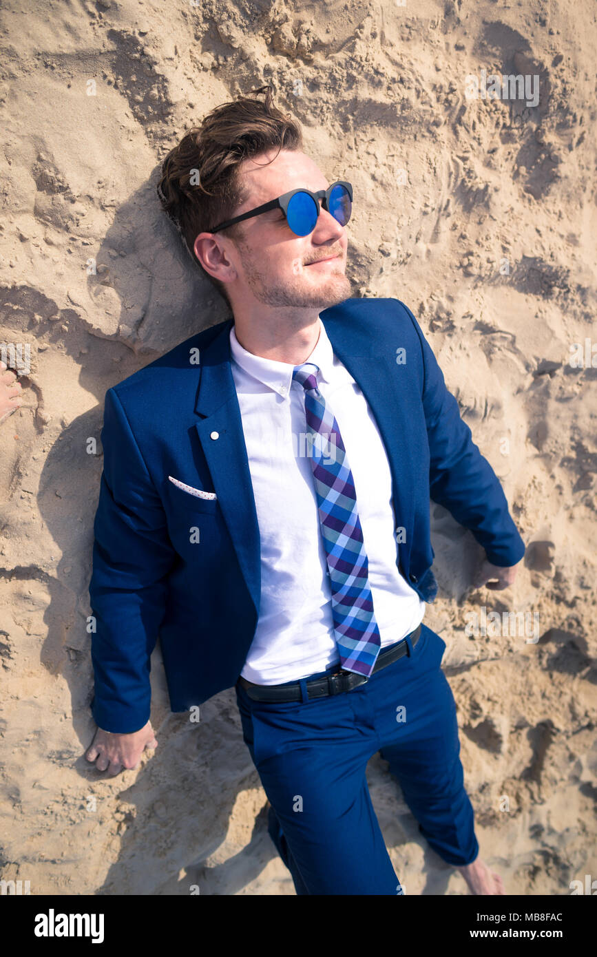 Lustig junge Mann im eleganten Anzug liegt am Strand. Stilvolle Kerl ist Sonnenbaden in den sonnigen Tag auf dem Sand. Gerne ungewöhnliche Manager bei Sonnenbrillen Stockfoto