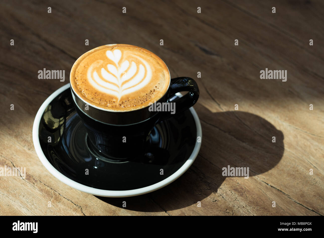 Close up Hot schwarz Kaffee Tasse mit Baum Herzform latte Art Milch Schaum auf Holz Tisch am Fenster mit harten Schatten Sonnenlicht im Cafe Restaurant. Stockfoto