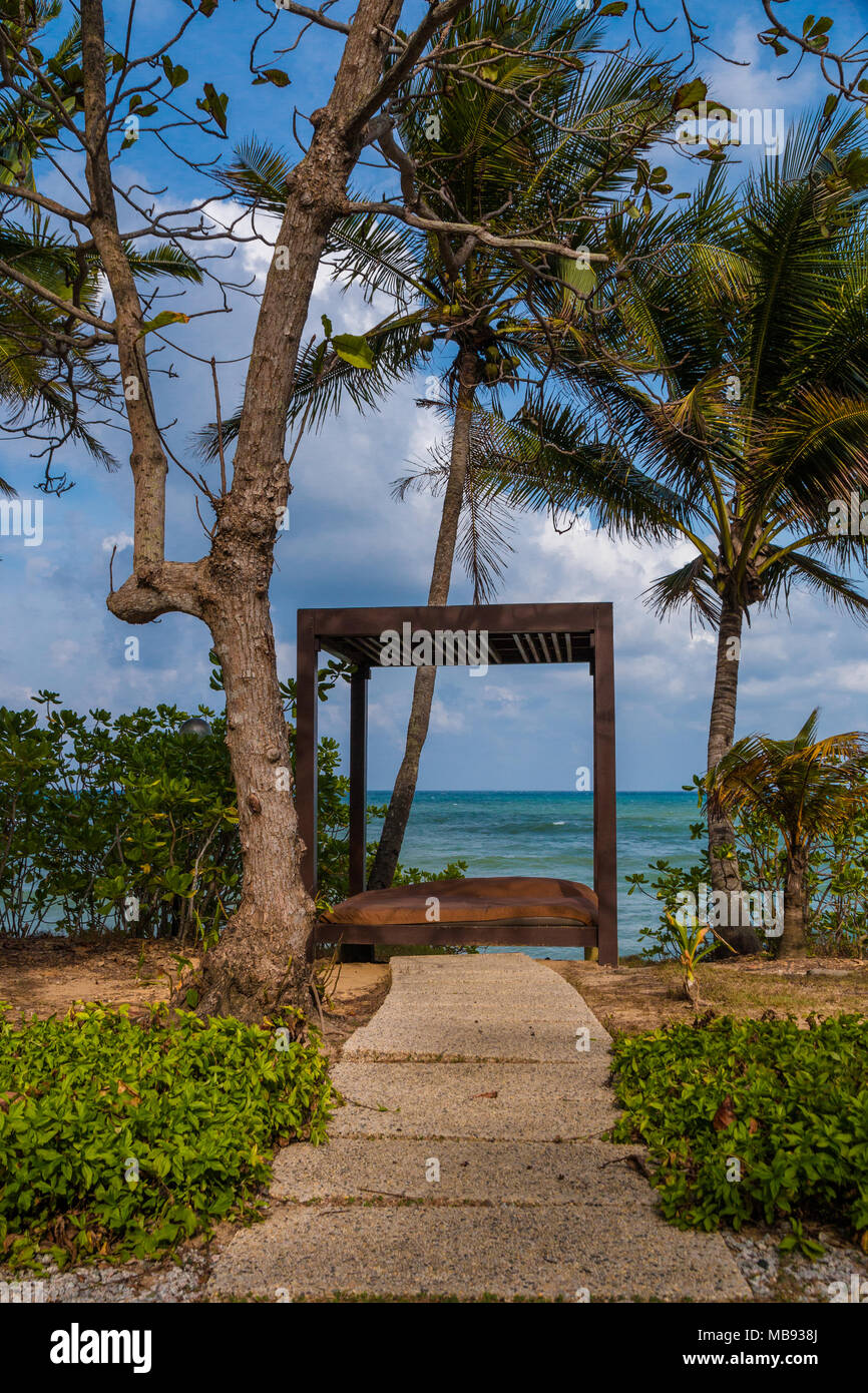 Ein Weg in eine offene Holz- Strand Cabana mit einer Matratze im Inneren, von grünen Pflanzen und Palmen umgeben, mit Blick auf einen wunderschönen Blick auf das Meer. Stockfoto