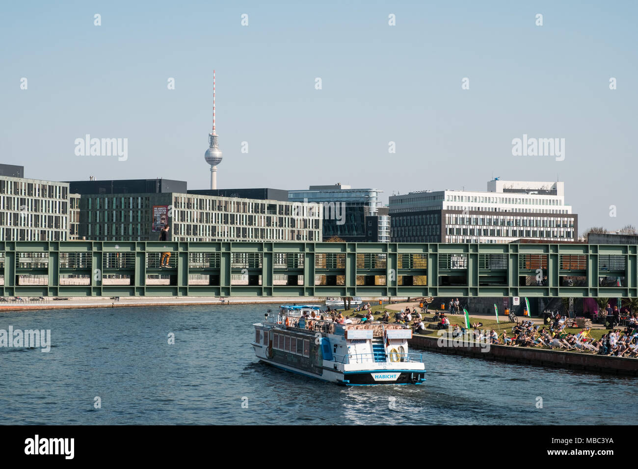 Berlin, Deutschland - April 2018: Berliner Stadtbild mit Schiff auf Spree, Leute sitzen auf Riverside und Fernsehturm im Hintergrund Stockfoto