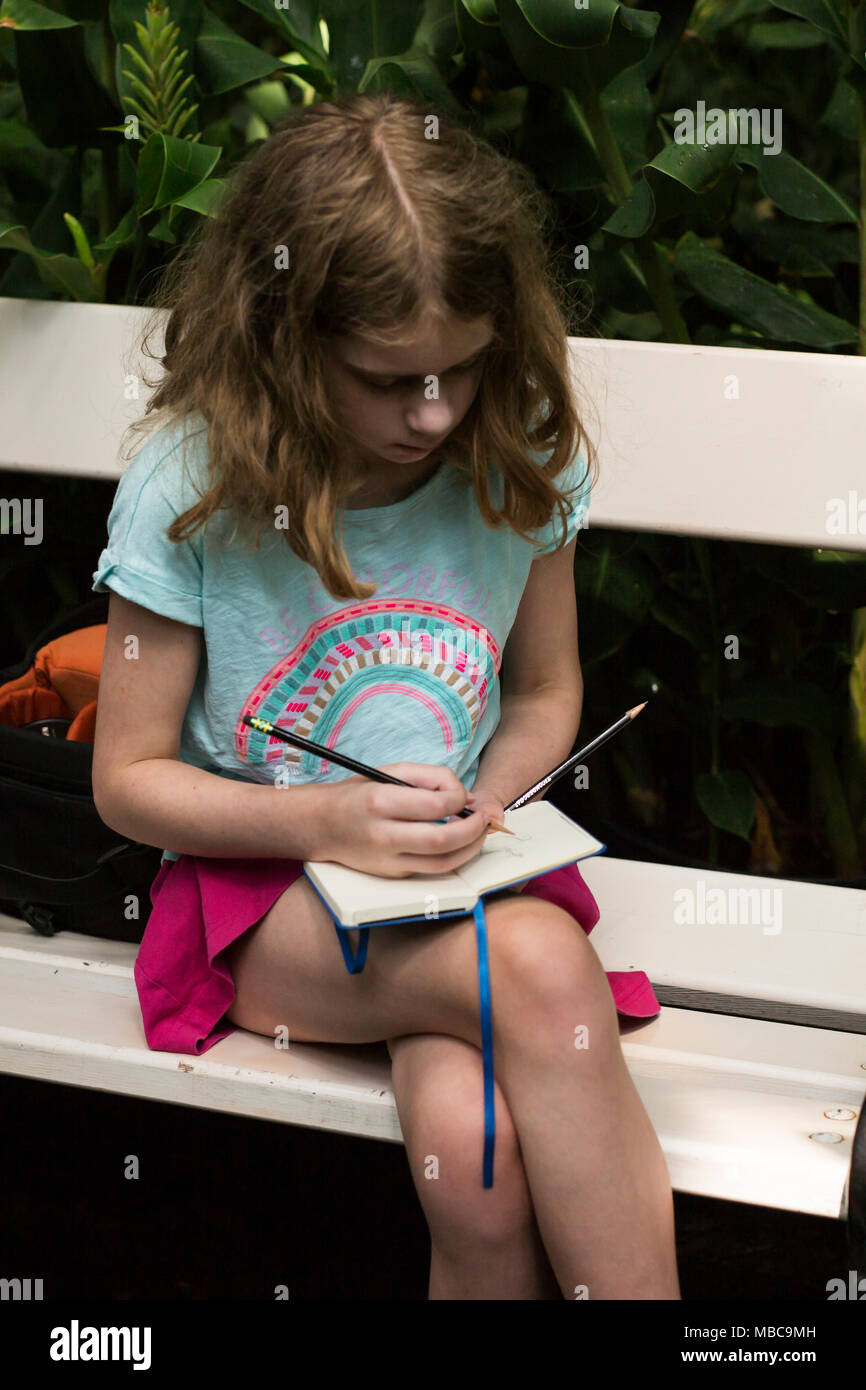 Ein zehn Jahre altes Mädchen sitzen auf einer Bank, Zeichnung Blumen in einem Gewächshaus. Stockfoto