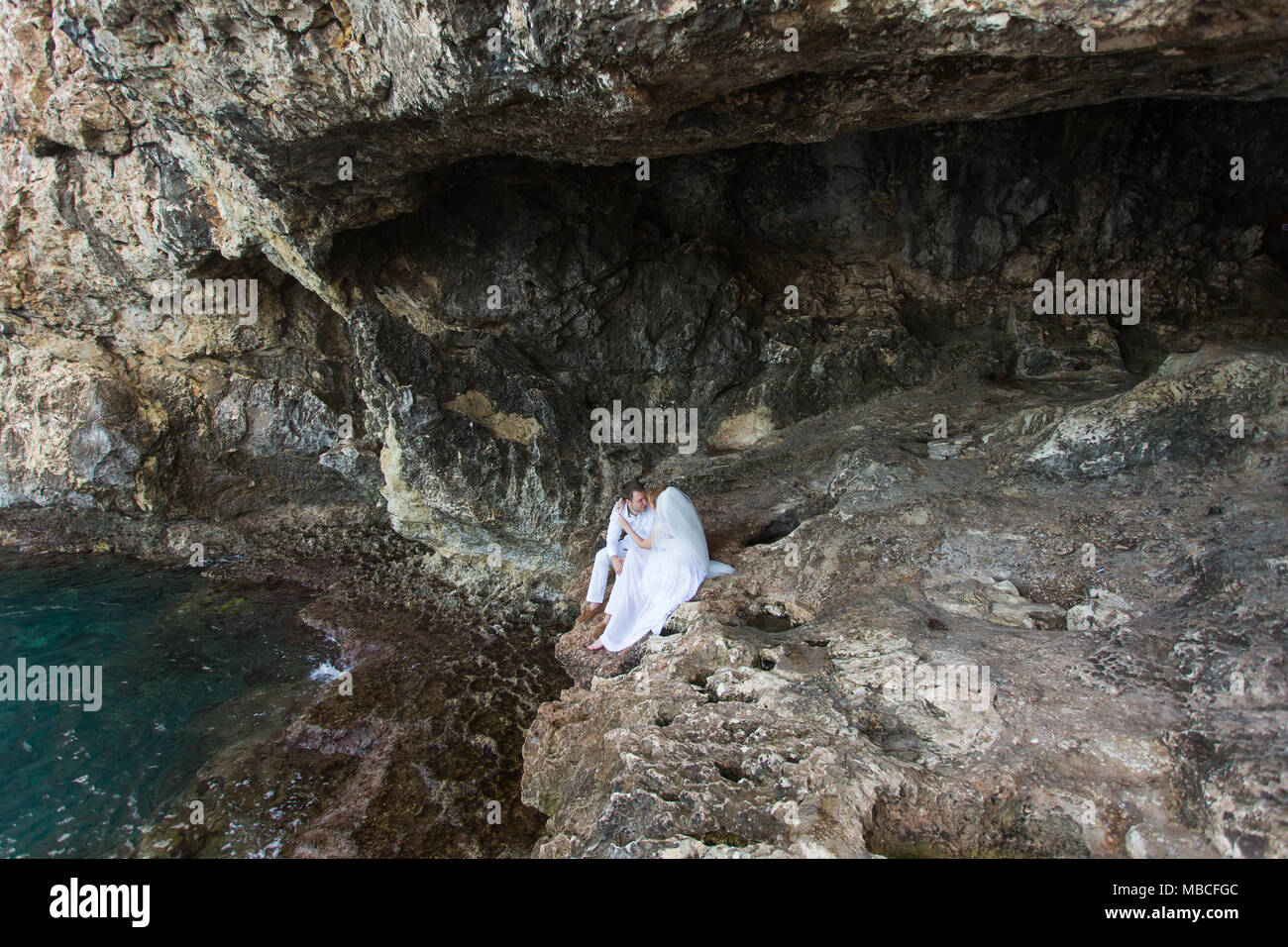 Paar Brautpaar Braut und Bräutigam Lachen und Lächeln zu einander, glücklichen und freudigen Moment. Der Mann und die Frau in der Hochzeit Kleidung sitzen auf dem Rock Hintergrund. Stockfoto