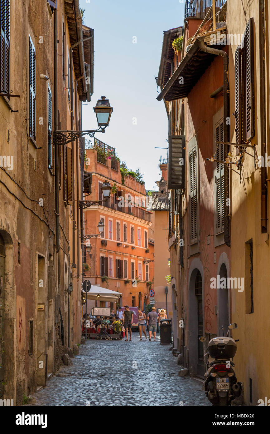 Eine Ikone der italienischen Szene mit Touristen entlang einer kopfsteingepflasterten Straße im Stadtteil Trastevere in Rom, Italien, vorbei an einem Restaurant mit Menschen außerhalb der Sat Stockfoto