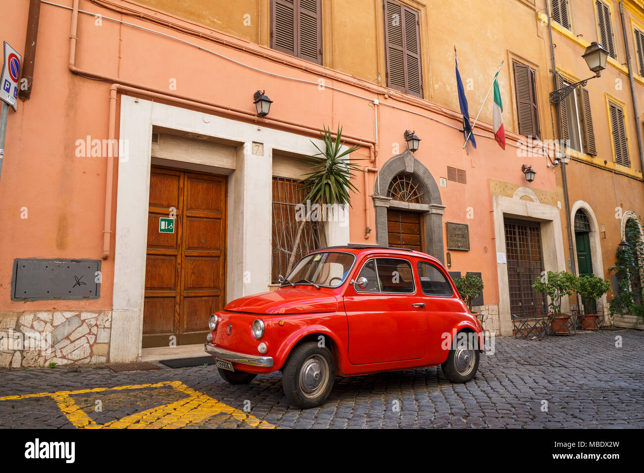 Iconic Ansicht einer alten roten Fiat 500 in einer gepflasterten Straße in Rom, Italien, geparkt, außerhalb eines Gebäudes unter italienischer Flagge. Die Nummernschilder zeigen ein MI Stockfoto