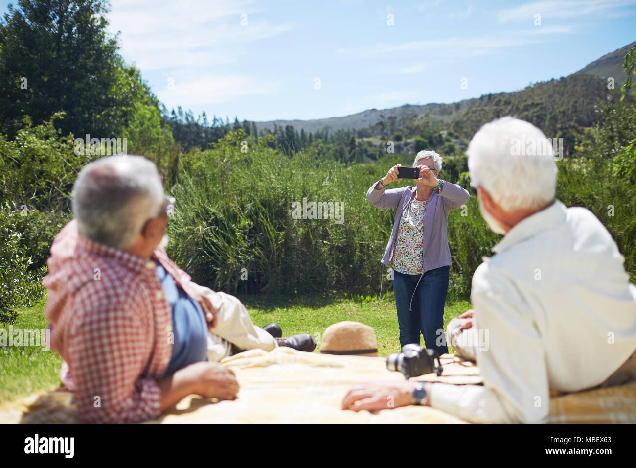 Active Senior Frau mit Kamera Handy fotografieren Männer entspannen auf sonnigen Sommer Picknickdecke Stockfoto