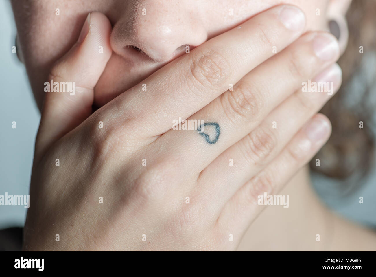 Eine Hispanic Frau deckt ihren Mund mit der Hand, in der Sie ein Tattoo von einem Herzen auf dem mittleren Finger sehen können Stockfoto