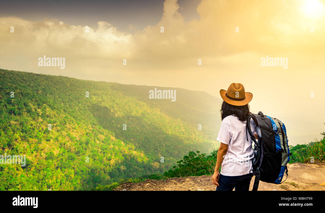 Junge Frau mit Rucksack unterwegs und Hut stehen oben auf der Klippe beobachten, schöner Blick auf Wald und Himmel nach Regen auf Ihren Urlaub Stockfoto
