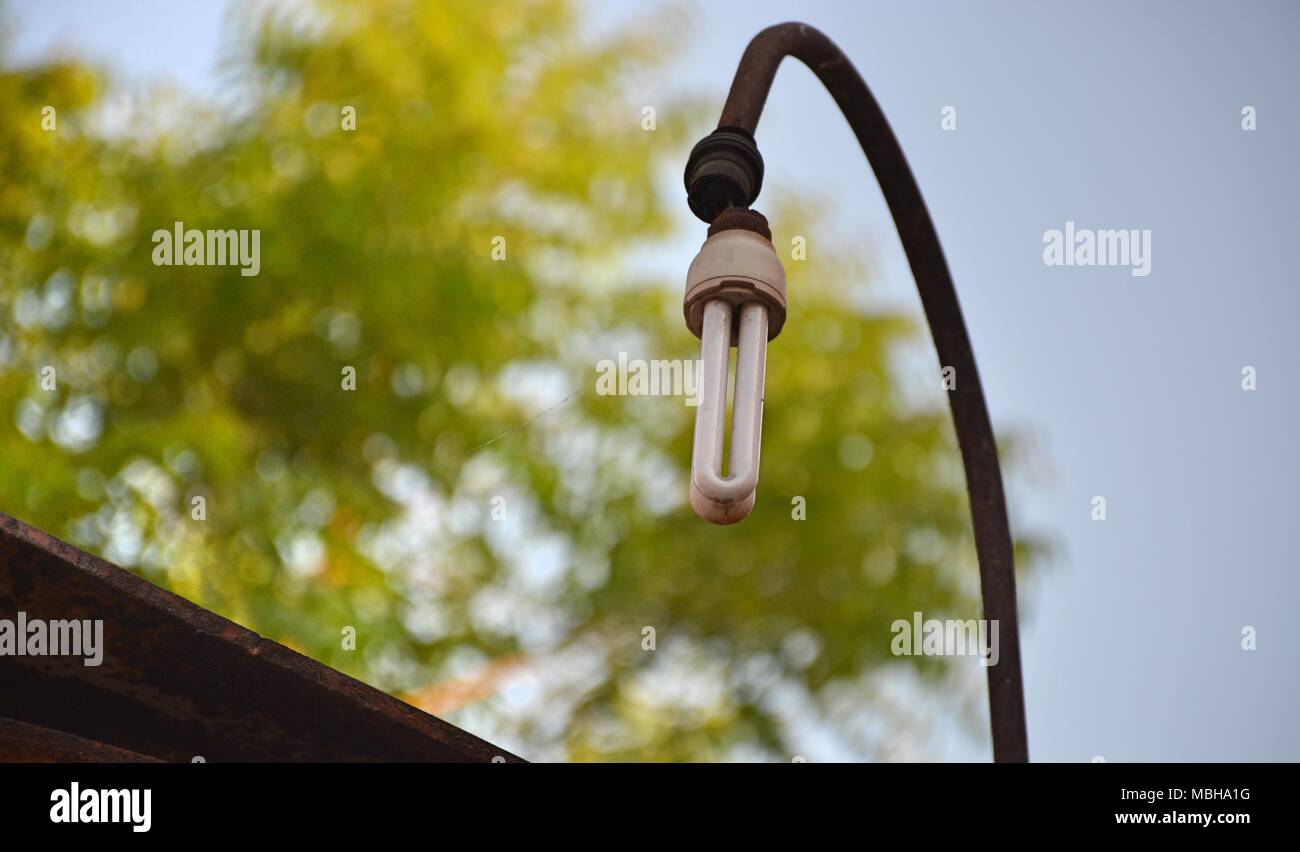 Beschädigte energy saver Glühbirne hängen mit einem korrodierte Lamp Post in der Öffentlichkeit mit Baum im Hintergrund Stockfoto