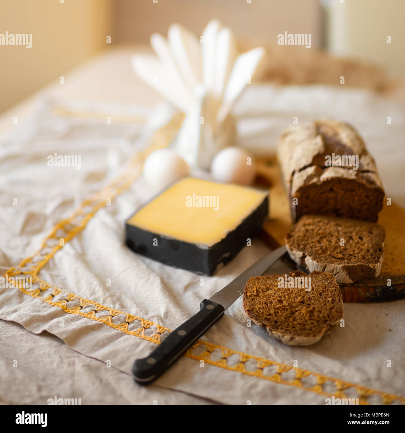 Ein Laib Brot mit in Scheiben geschnittenen Stücke auf einem Schneidebrett mit Messer, eine Scheibe Käse und Eier liegen auf einem Tisch mit einem Bettwäsche licht Tischdecke Stockfoto