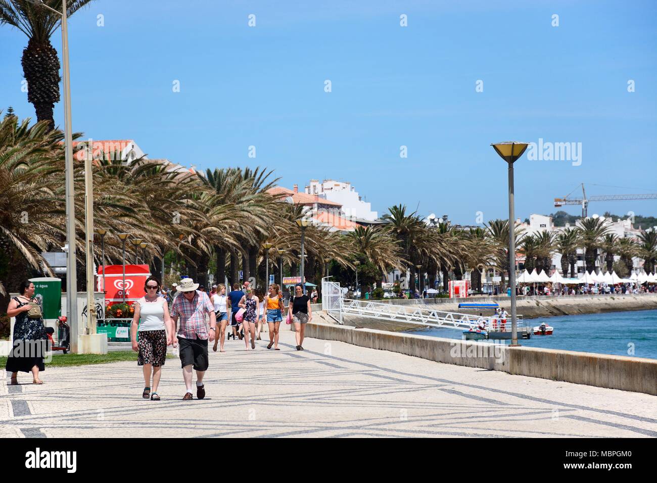 Touristen zu Fuß entlang der Palm Tree Avenida dos Descobrimentos mit Boote auf dem Fluss Bensafrim, Lagos, Algarve, Portugal, Europa gesäumt. Stockfoto