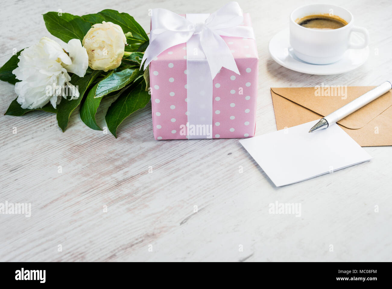 Pink gepunktete Geschenkbox, leere Grußkarte, Kraftpapier Umschlag, Pfingstrosen Bouquet und Kaffee Tasse über weißen Holzmöbeln rustikal Tisch. Romantischen Urlaub und Fall c Stockfoto