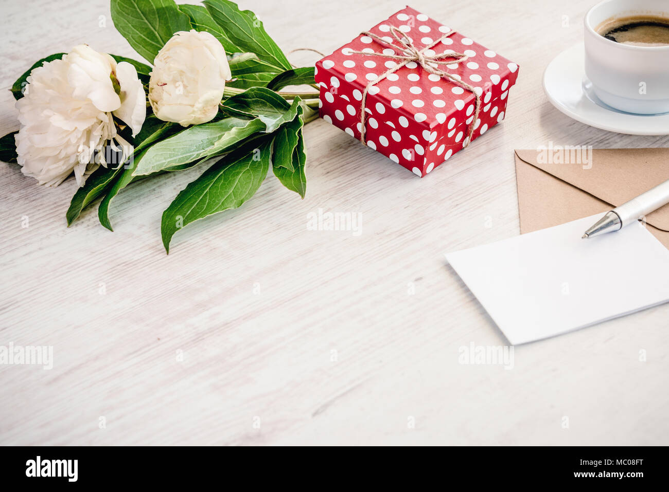 Gepunktete rote Geschenkbox, Kraftpapier Umschlag und leere Grußkarte, Kaffeetasse und Pfingstrose blumen blumenstrauss über weiß Holz Hintergrund. Kopieren Sie Platz. Stockfoto