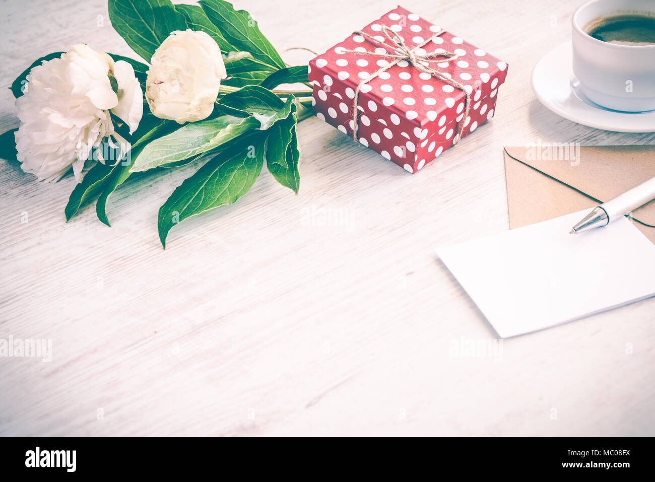 Gepunktete rote Geschenkbox, Kraftpapier Umschlag und leere Grußkarte, Kaffeetasse und Pfingstrose blumen blumenstrauss über weiß Holz Hintergrund. Kopieren Sie Platz. Vintage Effe Stockfoto